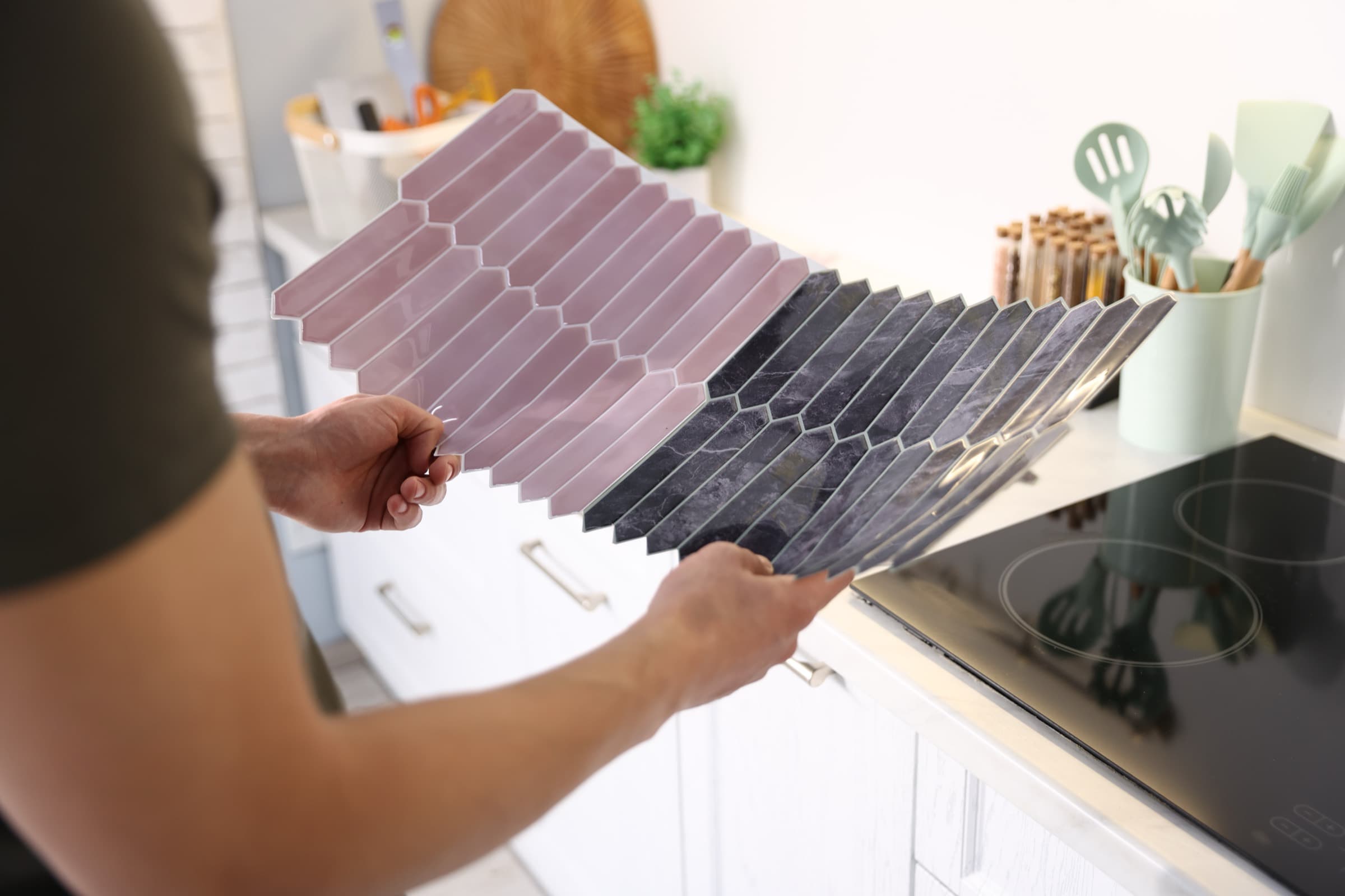 Hands holding pink and dark-marble peel-and-stick backsplash tile samples over a kitchen counter.